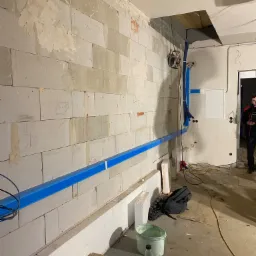 Wnętrze w trakcie remontu z widocznymi nieotynkowanymi ścianami z betonu komórkowego, niebieskimi rurami instalacji rozprowadzonymi wzdłuż ścian oraz instalacją elektryczną. Na podłodze widoczne...