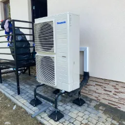 Zewnętrzna jednostka pompy ciepła Panasonic zamontowana na metalowej konstrukcji na kostce brukowej, z widoczną rurą odprowadzającą skropliny i elementami instalacji przy ścianie budynku.