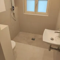 Jasna łazienka z beżowymi płytkami, kabiną prysznicową bez brodzika i umywalką na postumencie, okno z mleczną szybą.