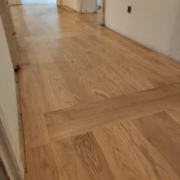 Ułożony parkiet z jasnego drewna w korytarzu, widoczne detale wzoru i łączenia desek, ściany w kolorze złamanej bieli, fragment ościeżnicy.