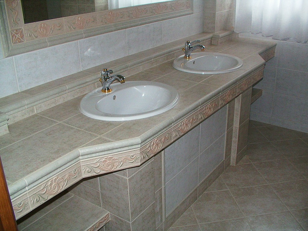 Umywalki ceramiczne nablatowe z chromowanymi bateriami, osadzone w blacie z płytek w odcieniach beżu, zdobionym ornamentami, z lustrem w tle.