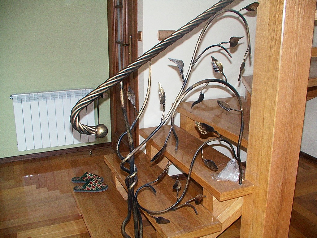 Drewniane schody z kutą, zdobioną balustradą w kształcie pnących się roślin, na podłodze leżą kapcie.