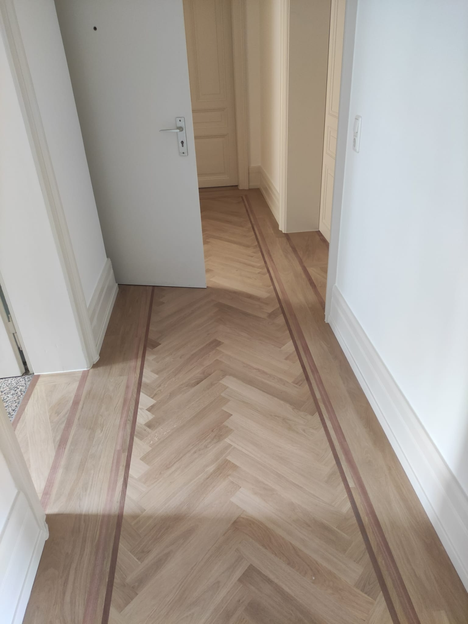 Ułożony parkiet w jodełkę z bordiurą z ciemniejszego drewna, widok na korytarz z białymi ścianami i otwartymi drzwiami.