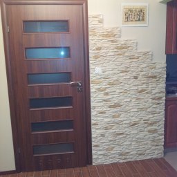 Gogobud - Ciemnobrązowe drzwi wewnętrzne z przeszkleniami, fragment dekoracyjnej ściany z imitacji kamienia ułożonej schodkowo, oraz podłoga z paneli imitujących drewno.