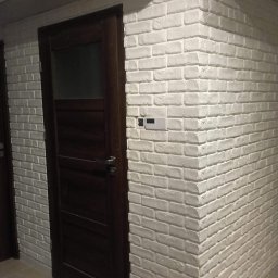 Gogobud - Ciemnobrązowe drzwi wewnętrzne z przeszkleniem, zamontowane w ścianie wyłożonej dekoracyjną białą cegłą, z widocznym włącznikiem światła.