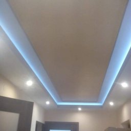 Gogobud - Beżowy sufit podwieszany z niebieskim oświetleniem LED wzdłuż krawędzi, widoczne drzwi z ciemnego drewna i wbudowane lampy punktowe.