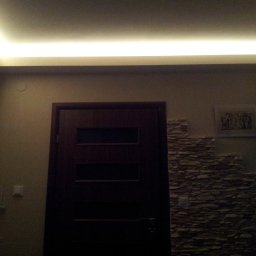 Gogobud - Ciemne drzwi wewnętrzne z szybkami, fragment ściany wykończony kamieniem dekoracyjnym i oświetlenie LED pod sufitem.