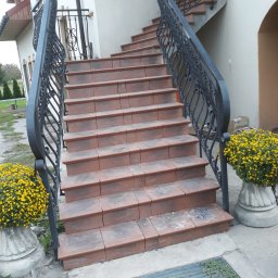 Gogobud - Zewnętrzne schody wyłożone terakotą z kutą, czarną balustradą, ozdobione donicami z żółtymi chryzantemami.