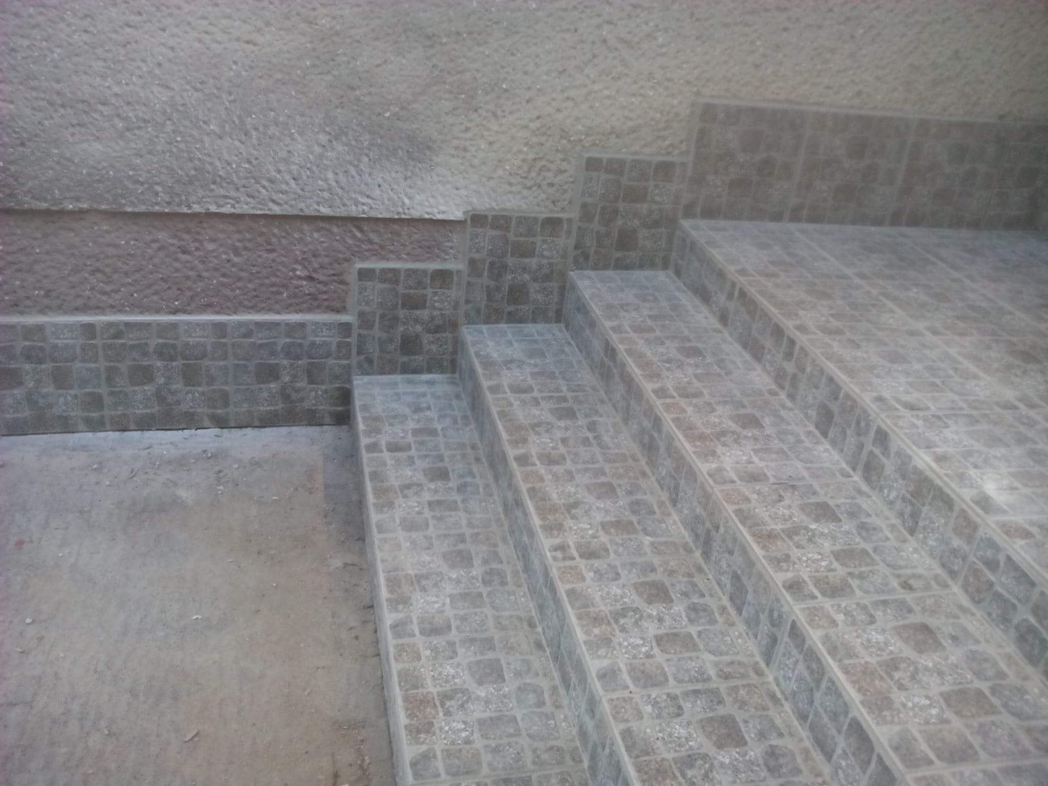 Wykończone płytkami schody zewnętrzne z szarej mozaiki, prowadzące do budynku z szarą i brązową elewacją, widok z dołu.