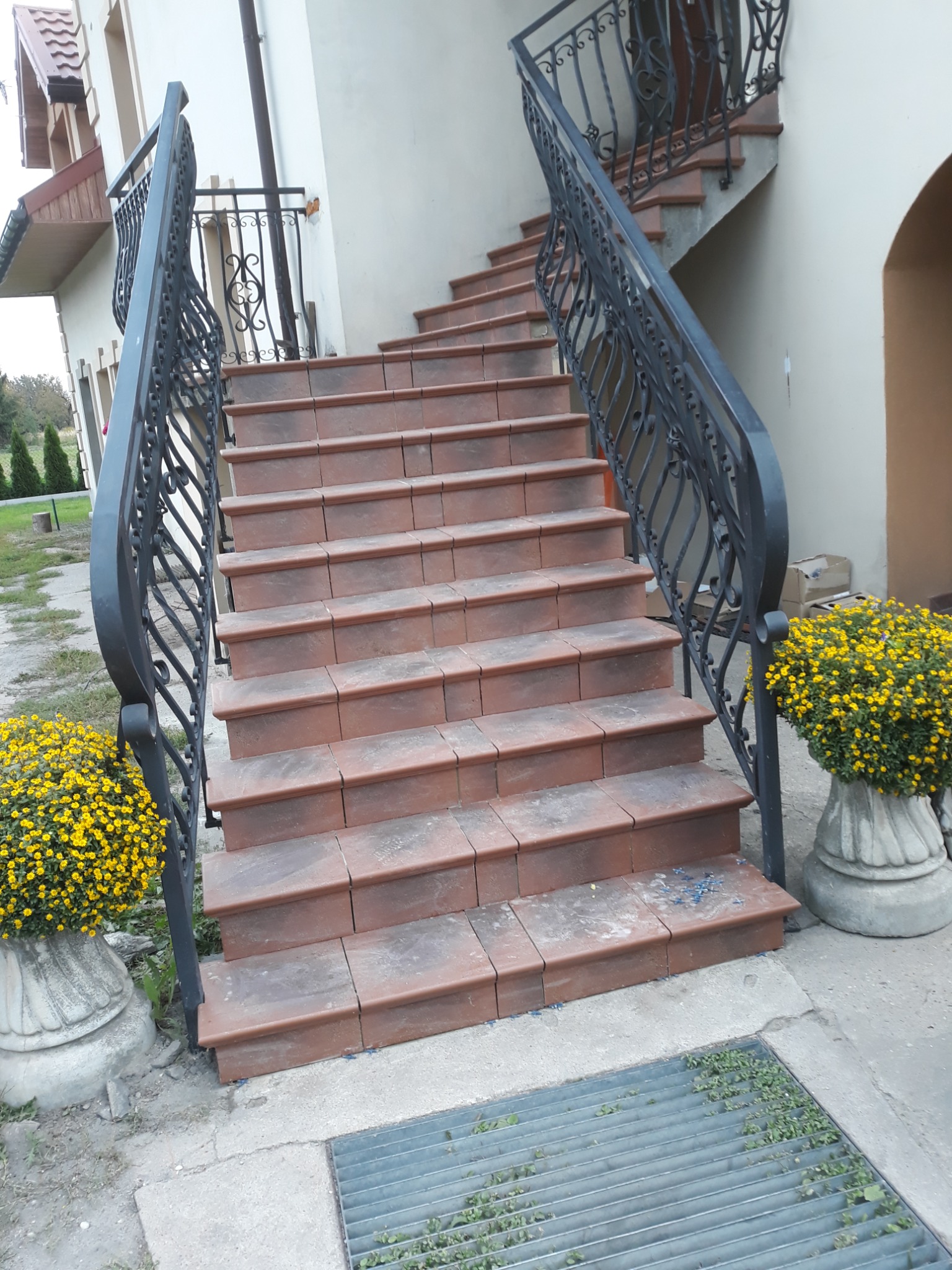 Zewnętrzne schody wyłożone terakotą z kutą, czarną balustradą, ozdobione donicami z żółtymi chryzantemami.