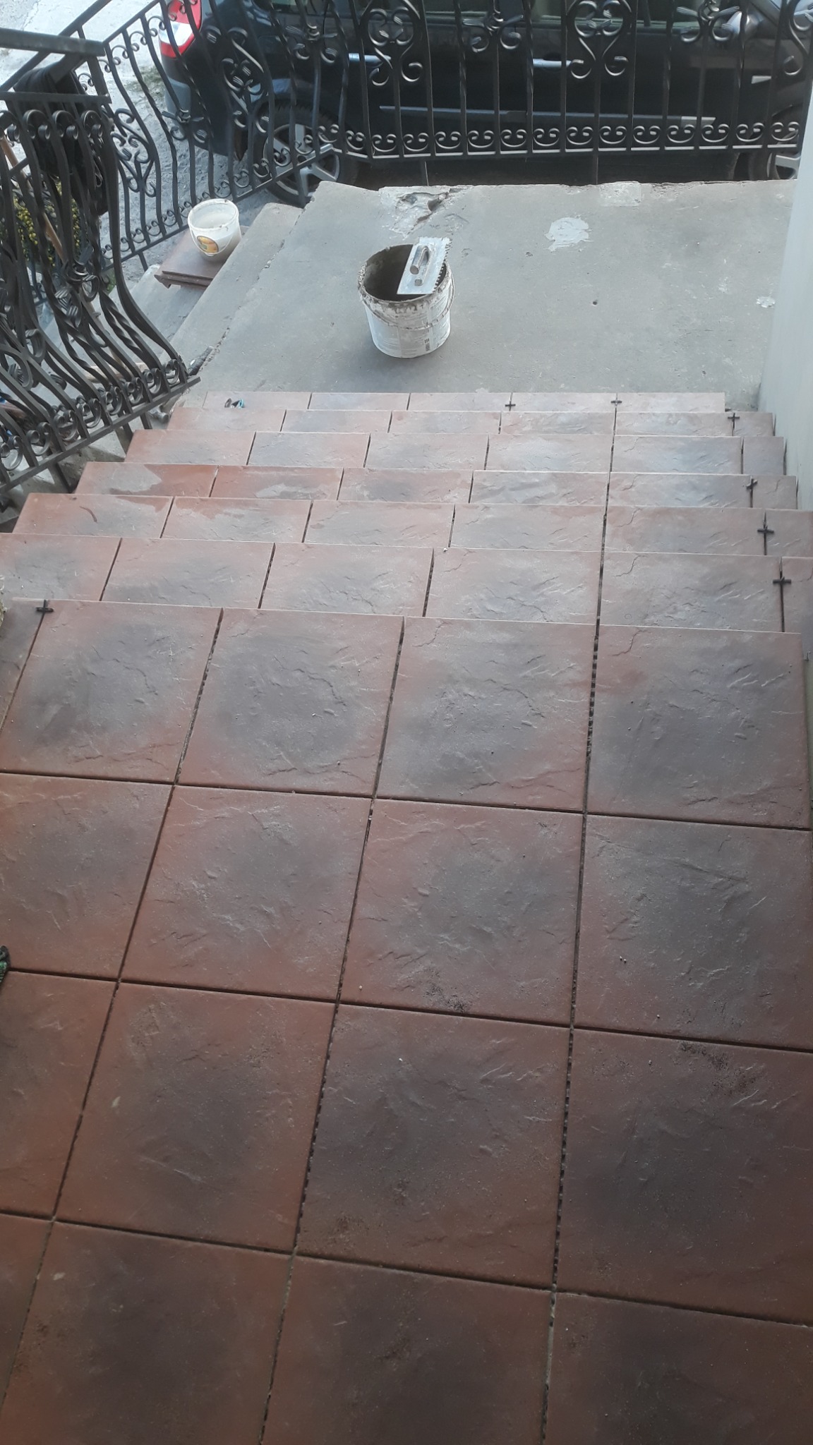 Kładzenie brązowych płytek na schodach zewnętrznych, widoczny kubeł z zaprawą i kielnią na górnym podeście, czarna metalowa balustrada po lewej.