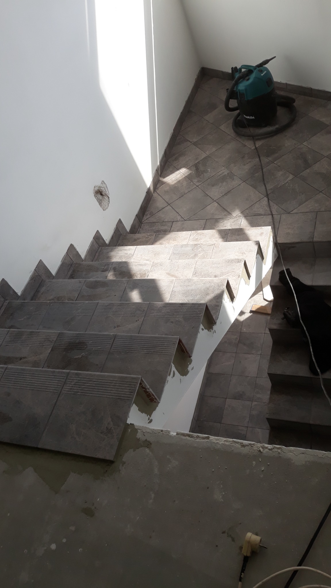 Schody w trakcie wykańczania, z szarymi płytkami na stopniach i podłodze, widoczny odkurzacz przemysłowy i przewody elektryczne, ślady zaprawy i niedokończone krawędzie.