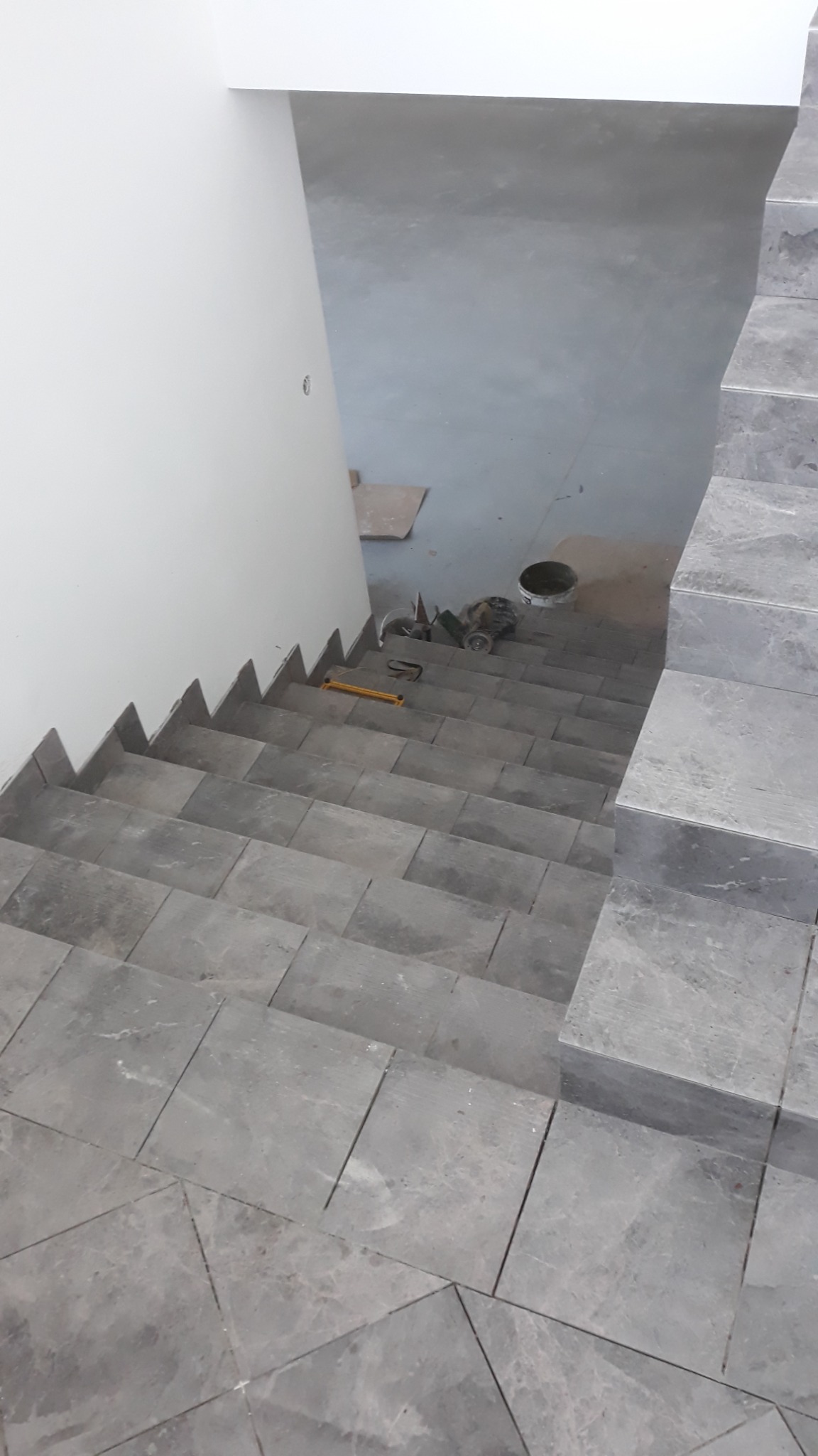 Układanie szarych, marmurkowych płytek na schodach w trakcie prac wykończeniowych w Grójcu; widoczne narzędzia glazurnicze i poziomica.