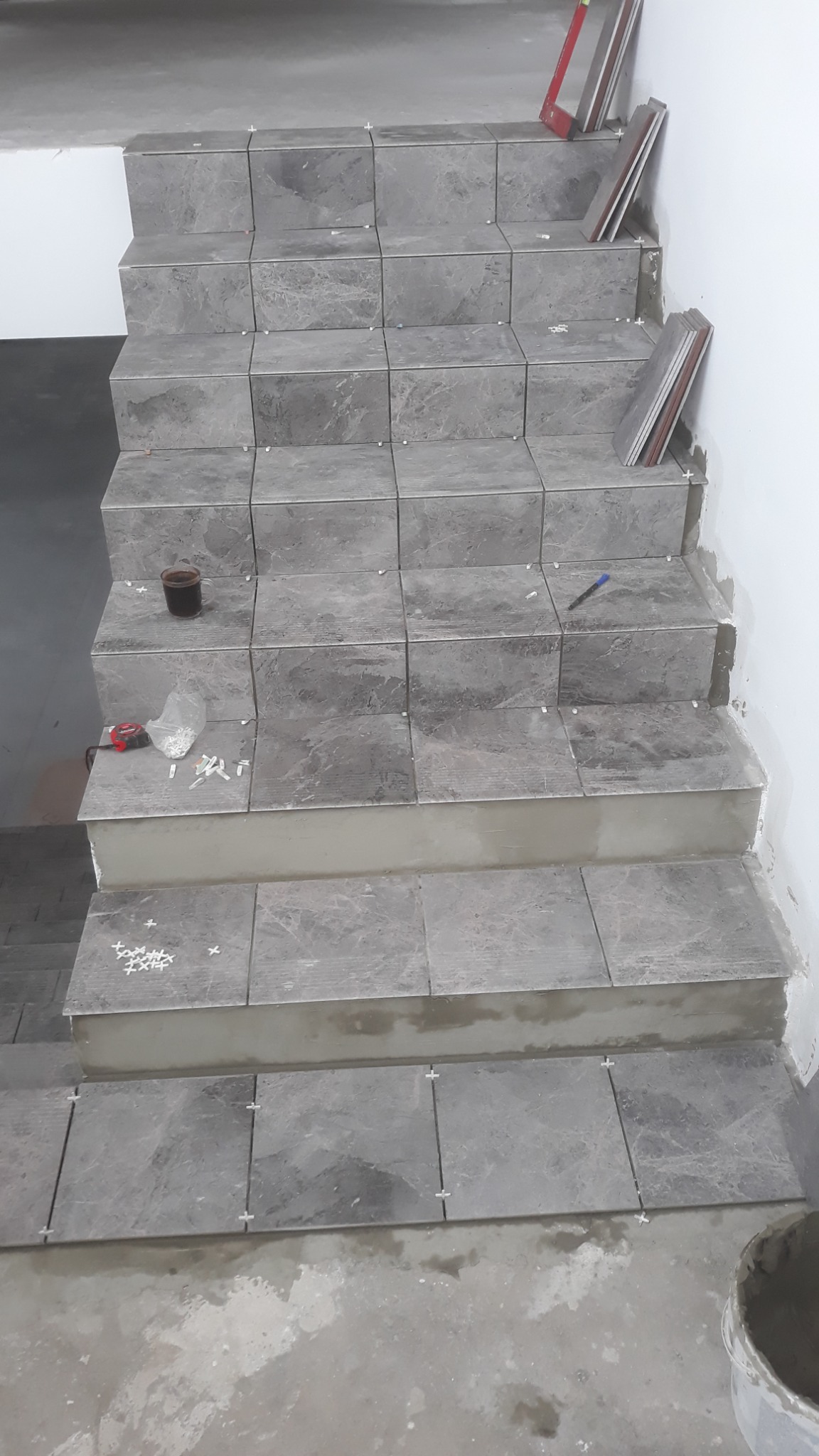Montaż płytek imitujących marmur na schodach wewnętrznych, widoczne krzyżyki dystansowe, poziomica i kubek z napojem.