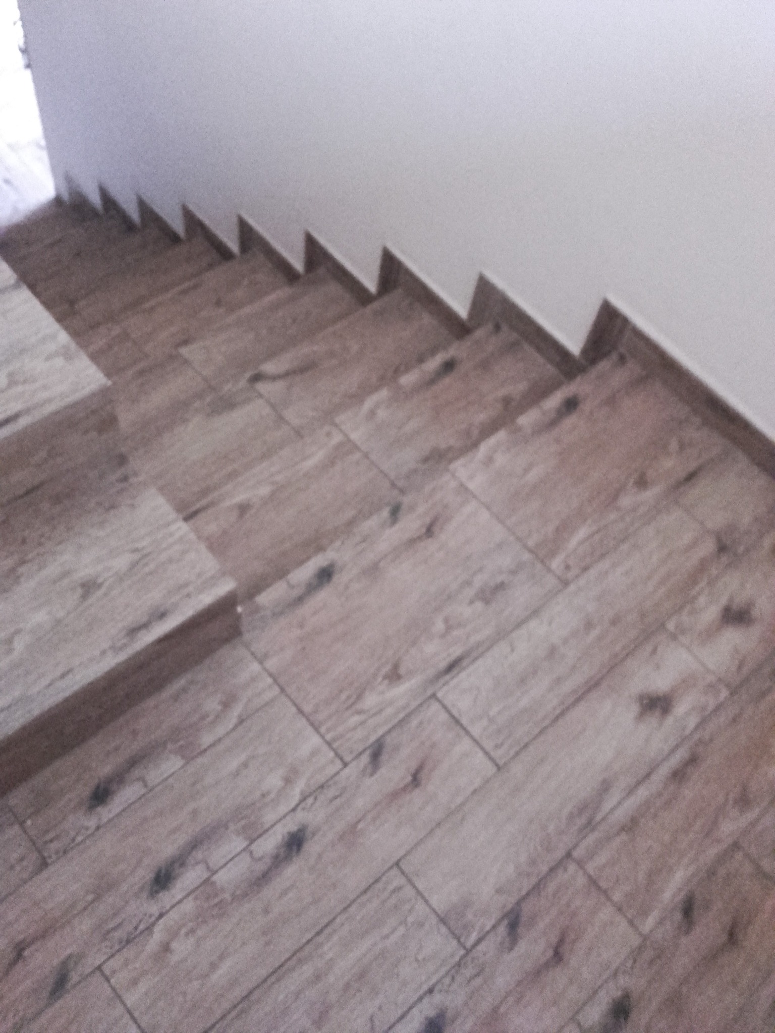 Klatka schodowa wyłożona płytkami imitującymi drewno, z widocznym wzorem słojów i naturalnymi przebarwieniami, prowadząca do góry, przy białej ścianie.