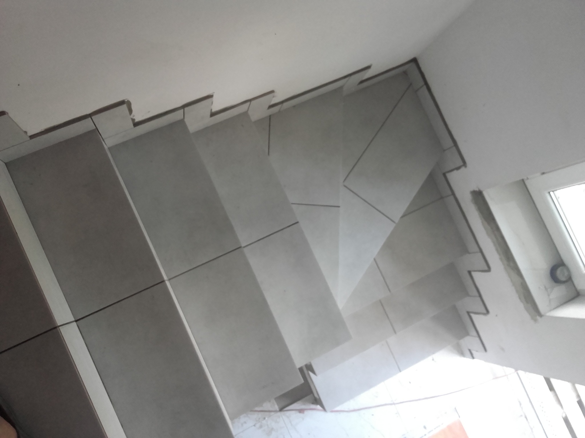 Widok z góry na schody wyłożone jasnoszarą, matową glazurą z ciemnymi fugami, widoczne niedociągnięcia przy krawędziach i okno w tle.