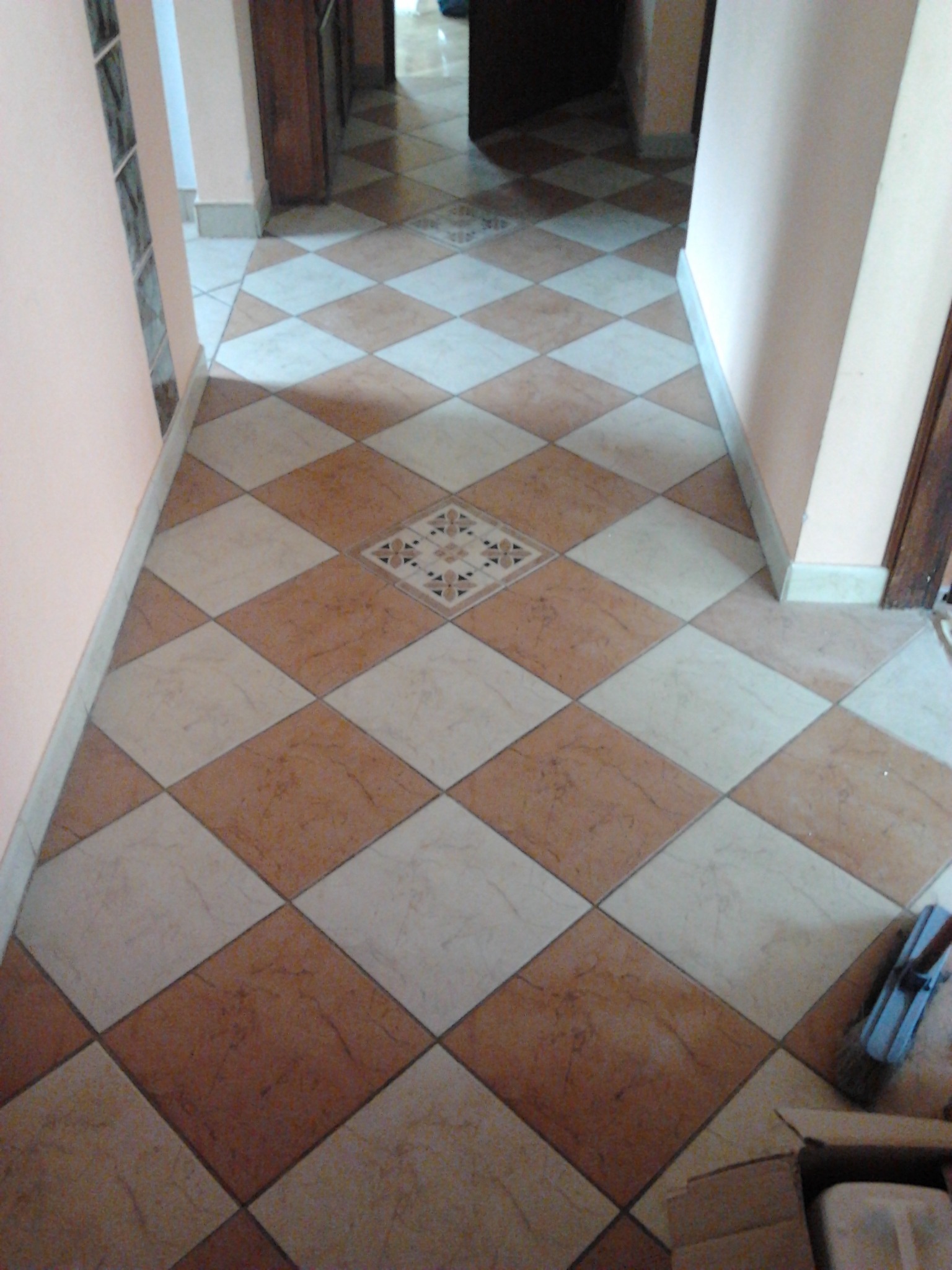 Ułożona podłoga z płytek w romby, beżowe i kremowe, z dekoracyjnym wzorem pośrodku i widoczną szczotką do zamiatania w rogu.