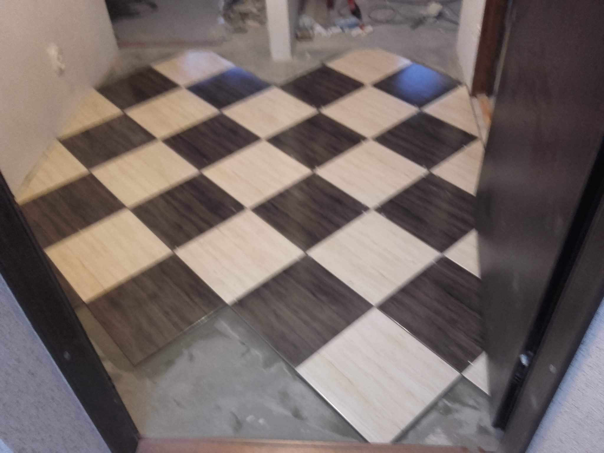 Układanie podłogi z płytek w szachownicę, widoczne niedociągnięcia w fugach i brak listew przypodłogowych, etap prac wykończeniowych.