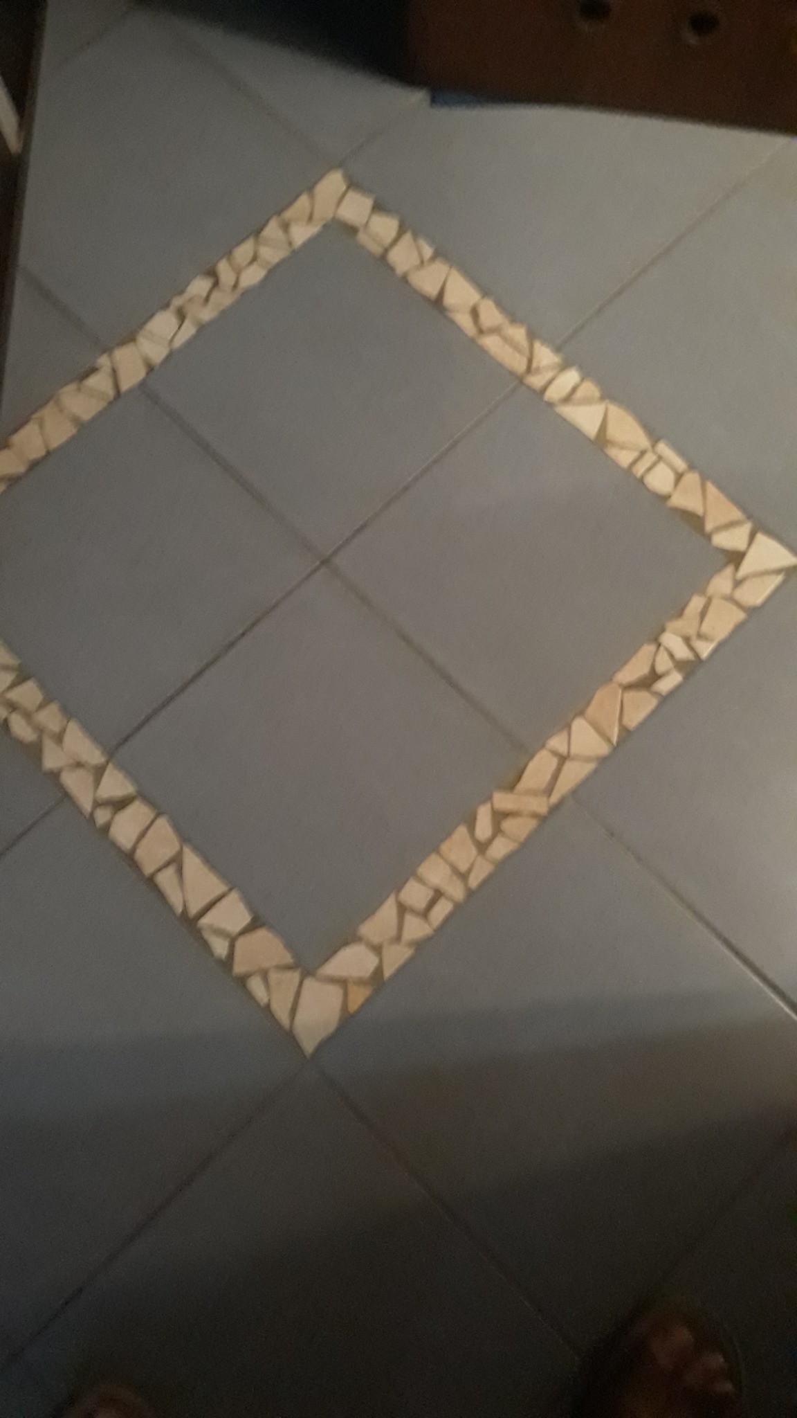 Podłoga wyłożona szarymi płytkami z dekoracyjnym obramowaniem w kształcie rombu z jasnych, nieregularnych kamyczków.