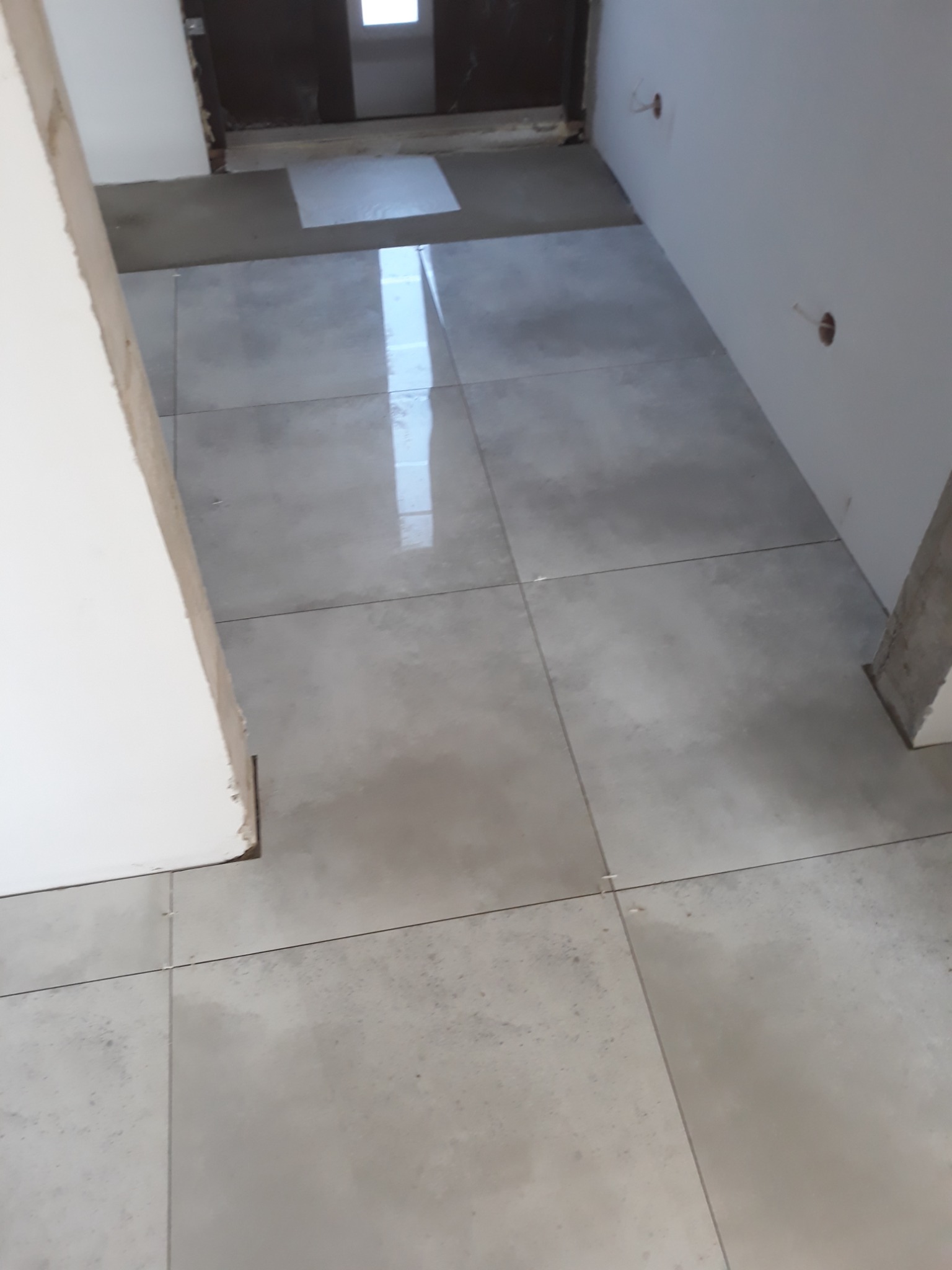 Świeżo ułożone, duże, szare płytki podłogowe w korytarzu, widoczne otwory na kable w ścianie, brak listew przypodłogowych, częściowo wykończone ościeżnice drzwi.