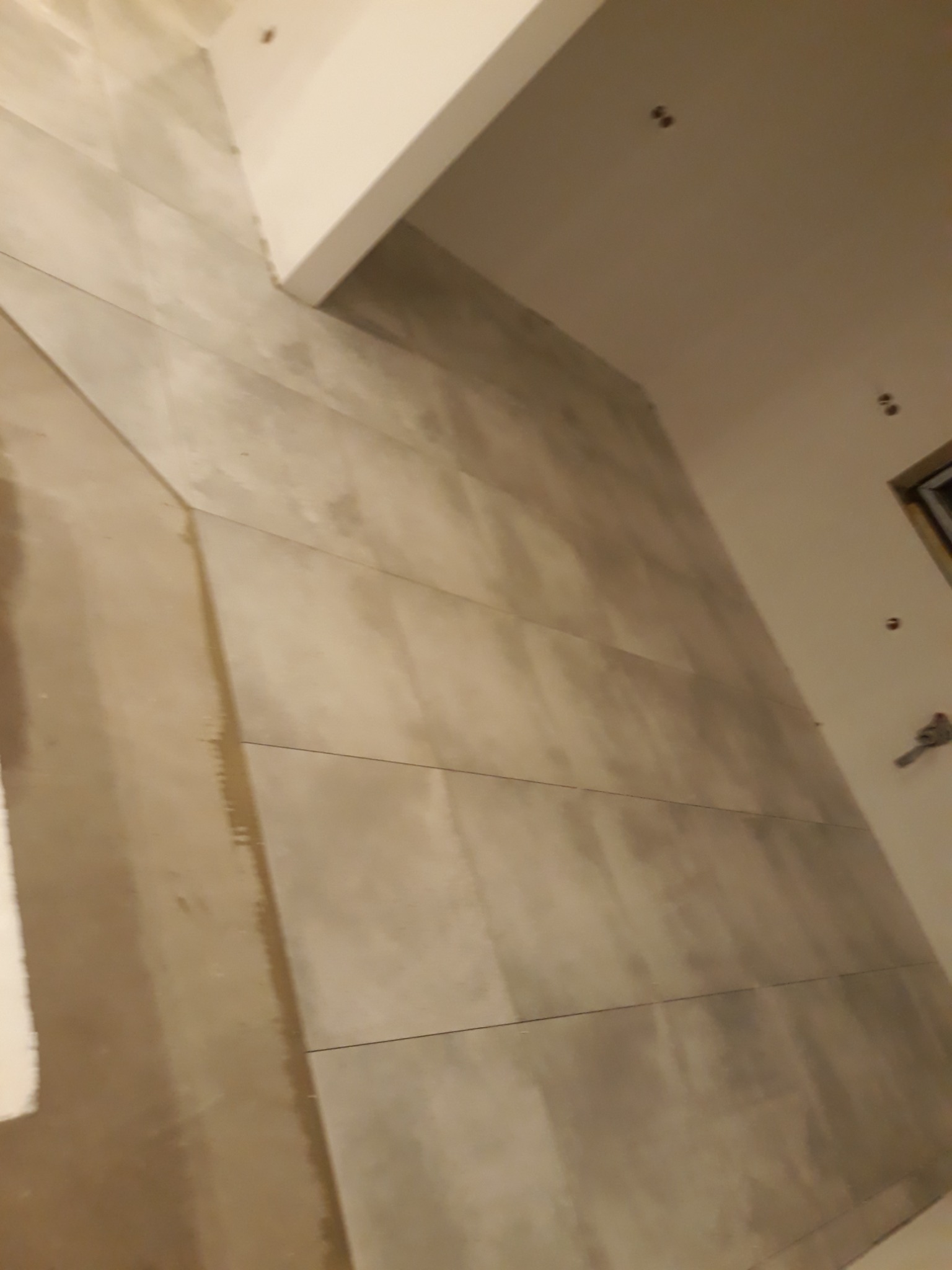 Ściana łazienki wyłożona dużymi, szarymi płytkami imitującymi beton, z widocznymi fugami i fragmentem sufitu z oświetleniem punktowym; w tle fragment okna i grzejnika.