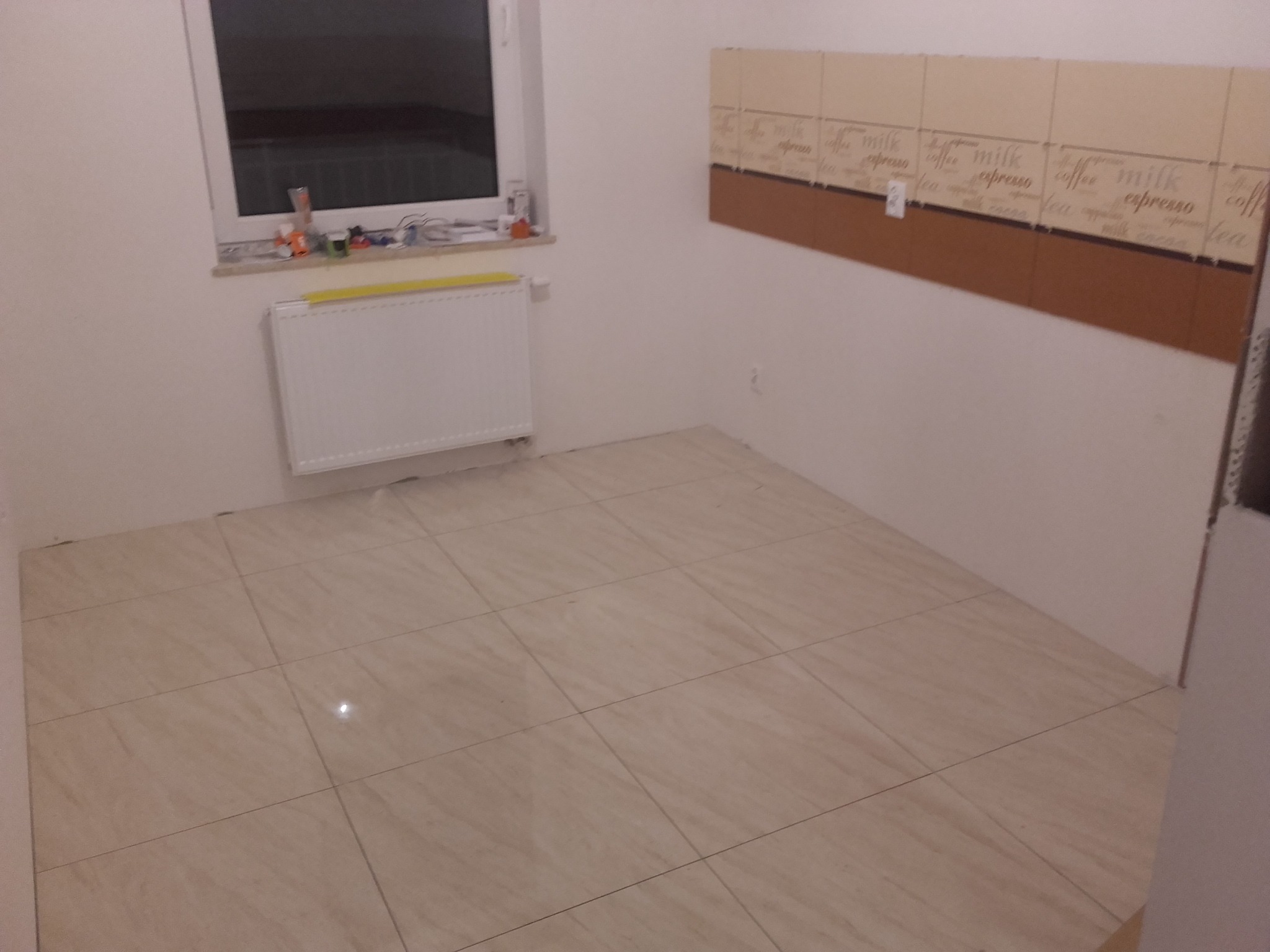 Świeżo ułożone, beżowe płytki podłogowe w kuchni, z fragmentem ściany z kremowymi i brązowymi kafelkami z motywem kawowym, biały grzejnik pod oknem.