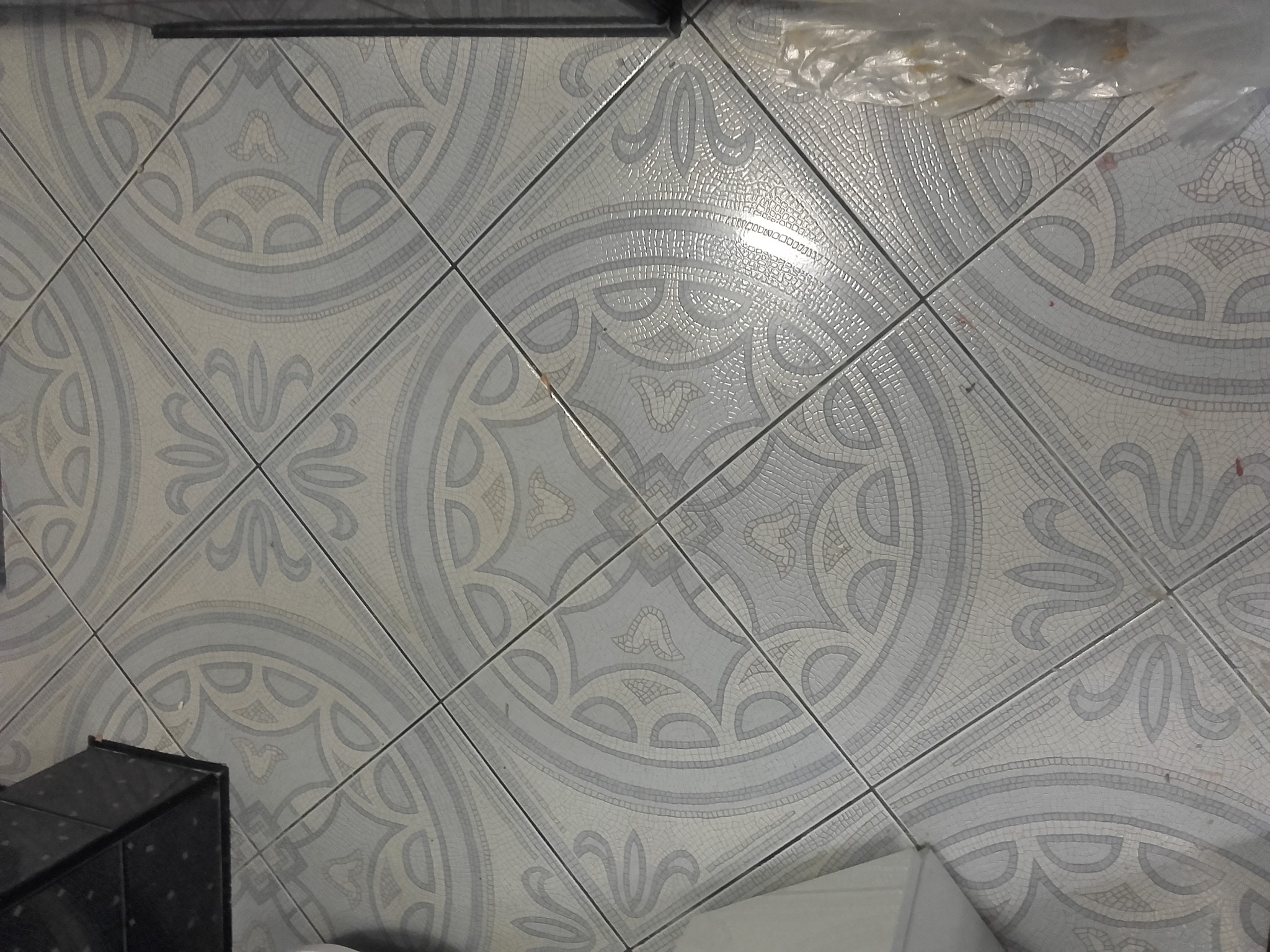Ułożone pod kątem, jasnoszare płytki podłogowe z wzorem kwiatowym i geometrycznym, częściowo zakryte folią ochronną i fragmentem czarnego mebla.