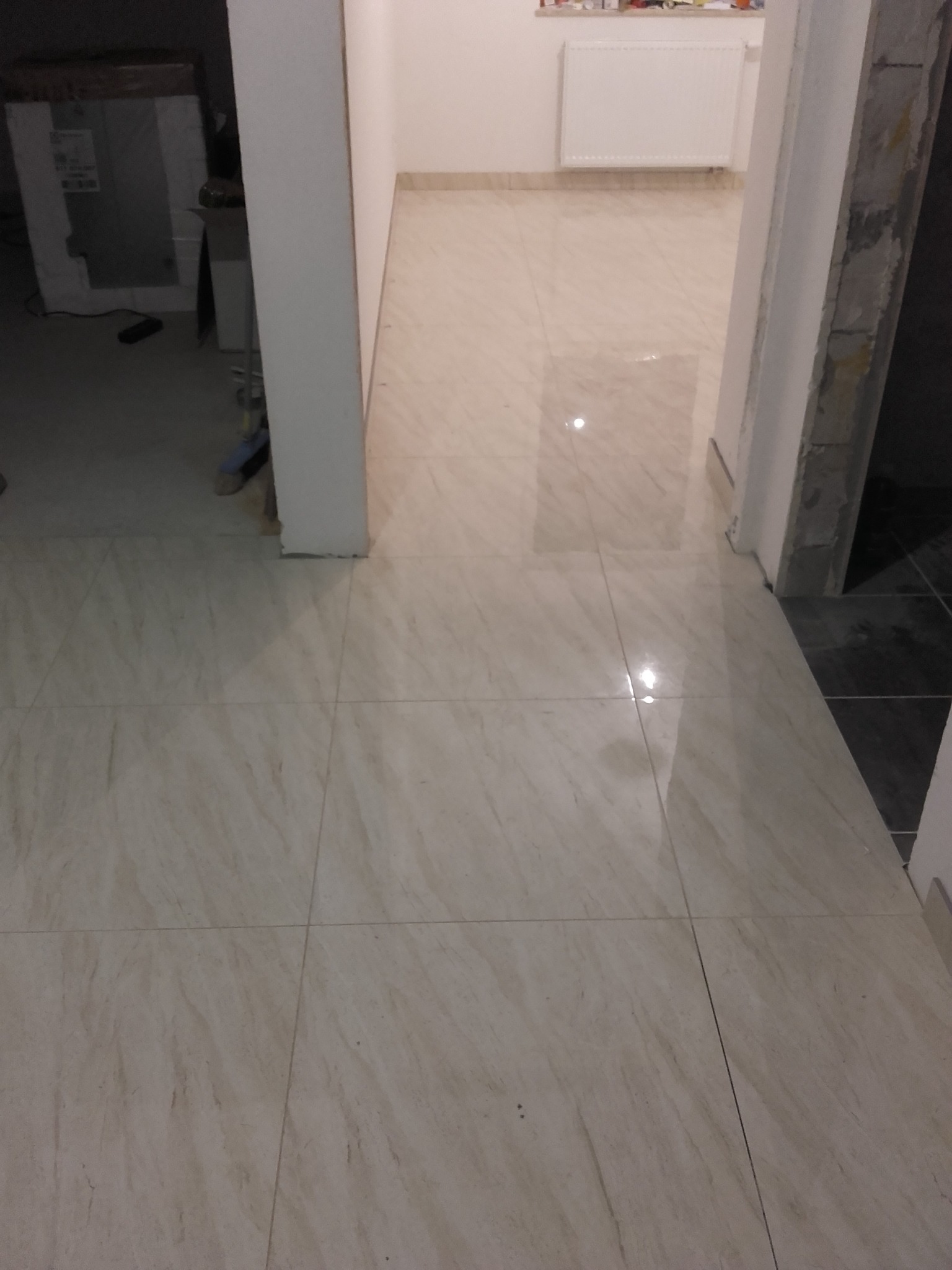 Połyskująca podłoga z płytek imitujących marmur w odcieniach beżu i kremu, świeżo położona w korytarzu, widoczne przejście do kolejnego pomieszczenia z grzejnikiem.