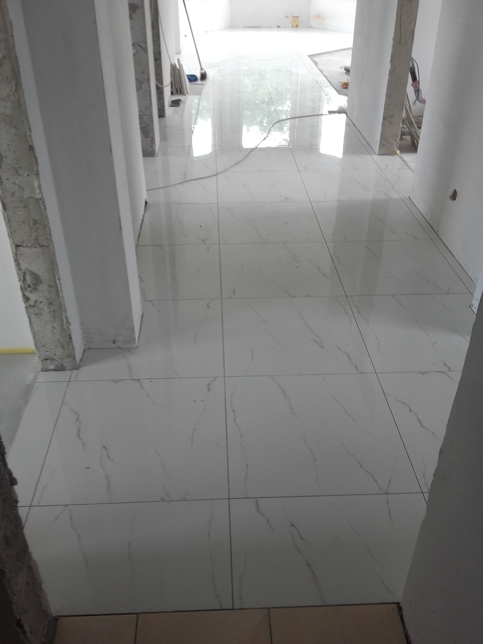 Białe, błyszczące płytki podłogowe z delikatnym marmurkowym wzorem w długim korytarzu w trakcie remontu, widoczne surowe ściany i kolumny.