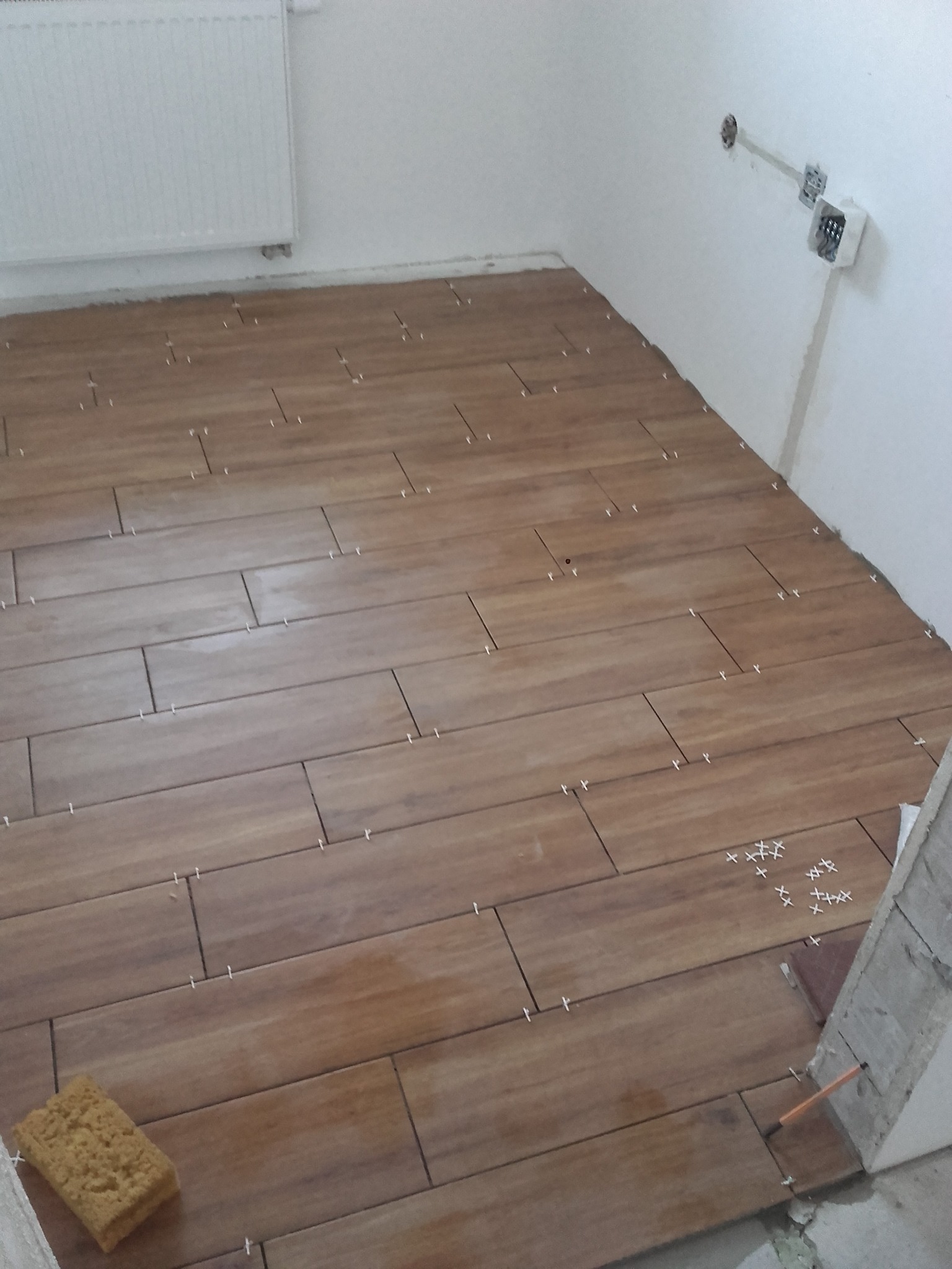 Układanie podłogi z płytek imitujących drewno w trakcie prac wykończeniowych, widoczne krzyżyki dystansowe, gąbka do czyszczenia i wystające kable elektryczne.