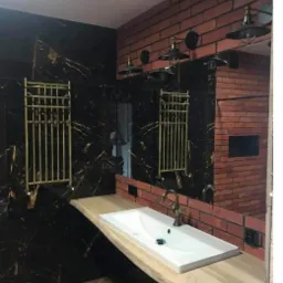 Nowoczesna łazienka z czarnymi płytkami imitującymi marmur, ceglaną ścianą, drewnianym blatem pod umywalkę i złotymi grzejnikami drabinkowymi.