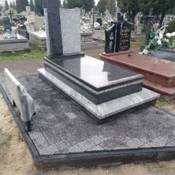 Nowoczesny, dwukolorowy pomnik z granitu w odcieniach szarości i czerni, umieszczony na wybrukowanym placu z kostki, z małą ławeczką zabezpieczoną folią, na tle innych nagrobków na cmentarzu.