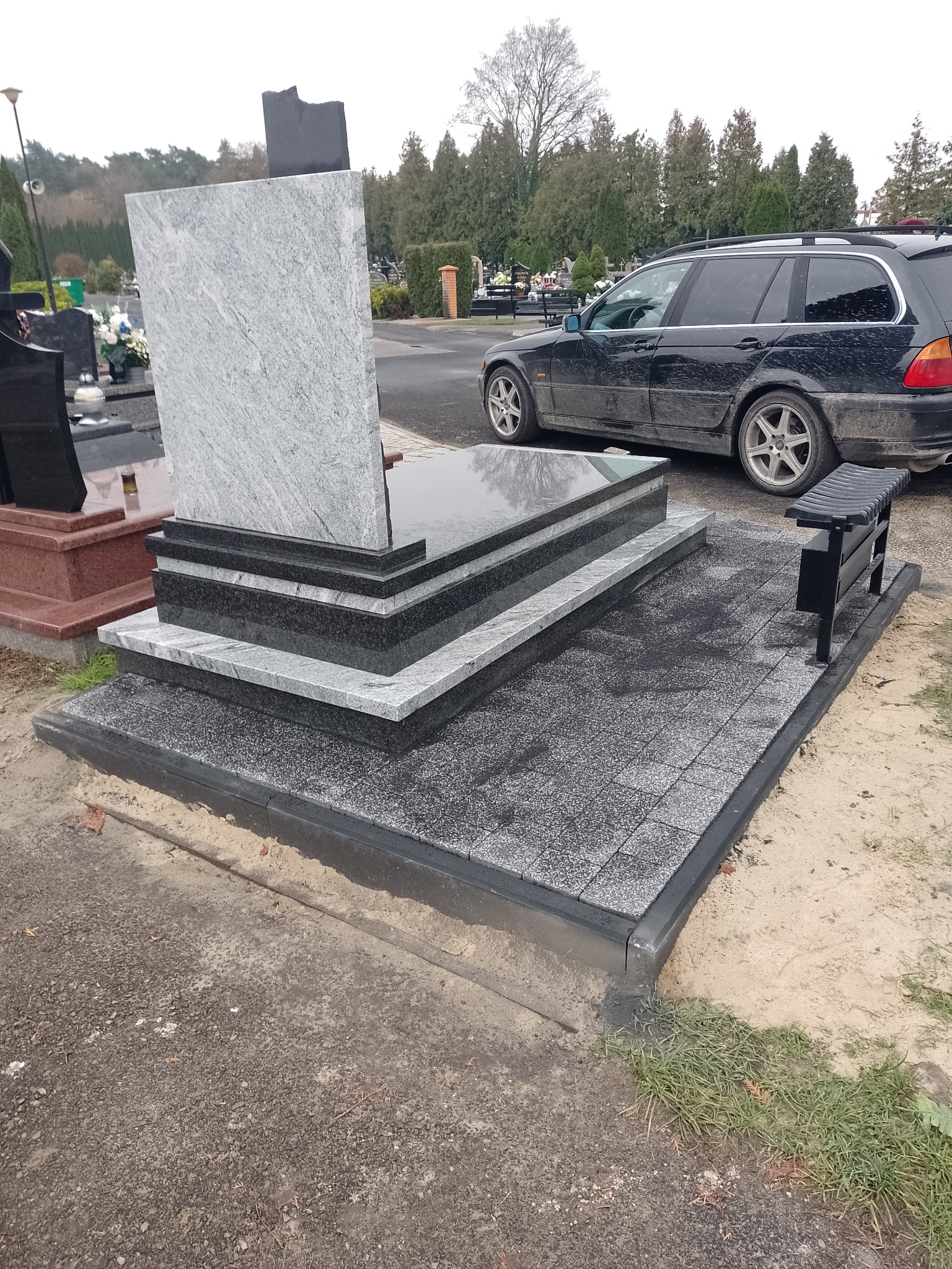 Granitowy nagrobek z jasnoszarą płytą i ciemnymi elementami, otoczony szarą kostką, z czarną ławeczką i samochodem w tle, na cmentarzu.