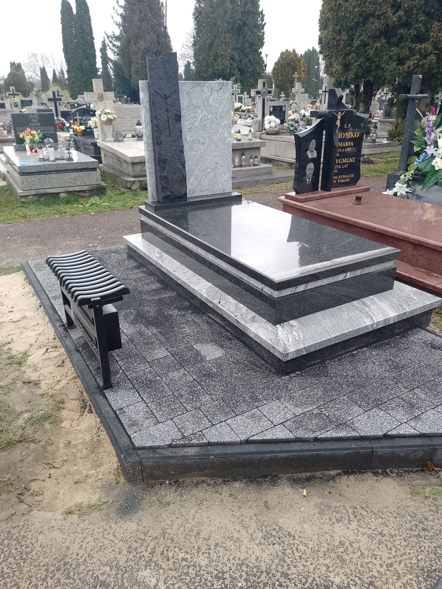Nowoczesny, dwukolorowy nagrobek z jasnego i ciemnego granitu, otoczony kostką brukową i wyposażony w czarną ławkę na cmentarzu.