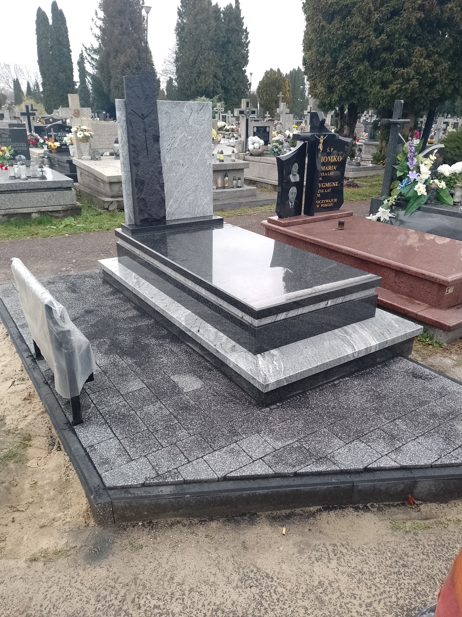 Nowoczesny, dwukolorowy pomnik z granitu w odcieniach szarości i czerni, umieszczony na wybrukowanym placu z kostki, z małą ławeczką zabezpieczoną folią, na tle innych nagrobków na cmentarzu.