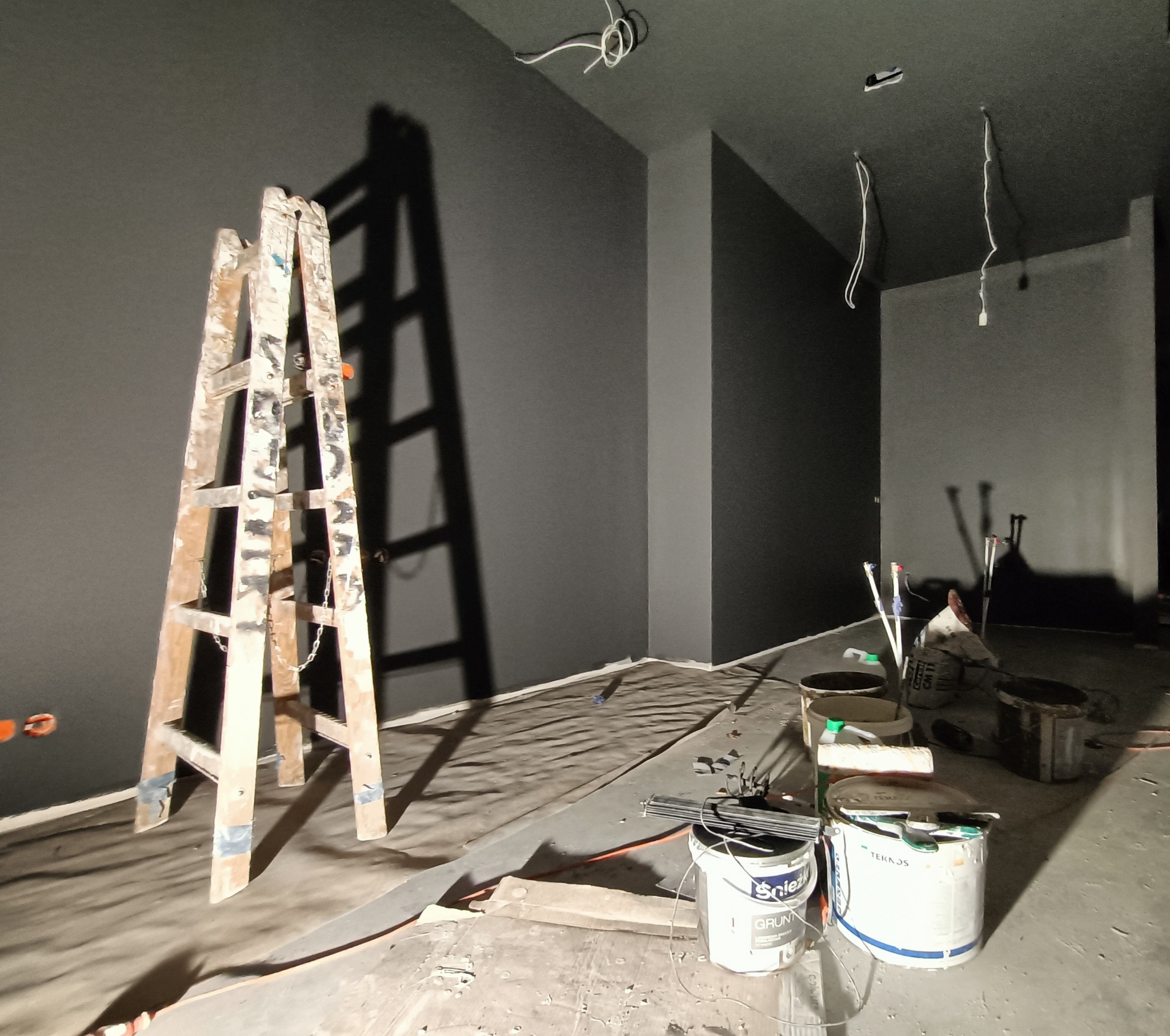 Wnętrze w trakcie malowania na ciemny kolor, widoczna drewniana drabina oparta o ścianę, puszki z farbą i narzędzia malarskie na podłodze zabezpieczonej folią malarską, wystające kable z sufitu.