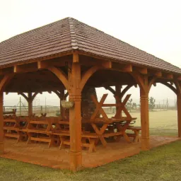 Drewniana altana z krytym dachem z dachówki ceramicznej, z drewnianymi stołami i ławami wewnątrz, osadzona na ceglanej podłodze, na zewnątrz widoczny trawnik i ogrodzenie.