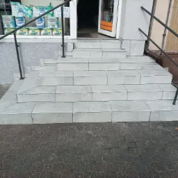 Wyłożone jasnoszarymi płytkami schody wejściowe do sklepu z metalową barierką, widoczne opakowania papieru toaletowego w witrynie.