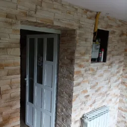 Ściana wykończona imitacją kamienia z widocznym licznikiem gazu, rurą gazową, grzejnikiem i drzwiami z naklejkami.