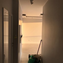 Malarz-Złota Rączka - Korytarz w trakcie odświeżania: świeżo pomalowane szare ściany, odnowiona podłoga z jasnych paneli, widoczne akcesoria malarskie w rogu, w głębi pokój z oświetleniem sufitowym.
