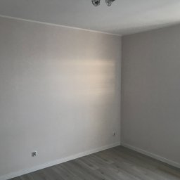 Malarz-Złota Rączka - Pomalowane na jasny odcień szarości ściany w rogu pokoju z drewnopodobną podłogą, widoczne dwa gniazdka elektryczne i grzejnik pod oknem.