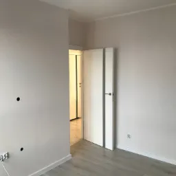 Świeżo pomalowane, jasnoszare ściany w pustym pokoju z otwartymi białymi drzwiami, widoczne gniazdka elektryczne i otwory w ścianie przygotowane do montażu osprzętu.