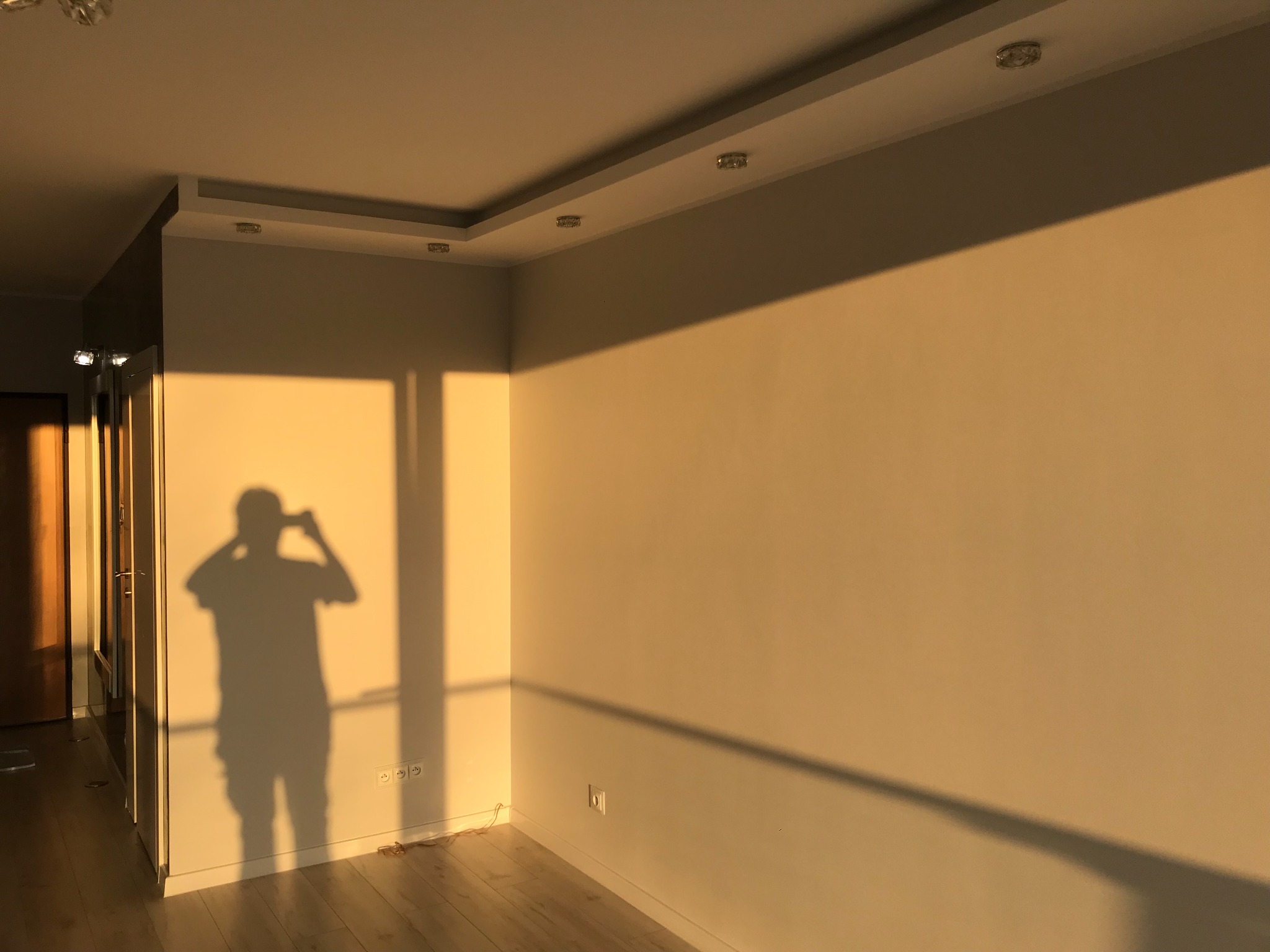 Pusty pokój z parkietem, oświetlony zachodzącym słońcem, rzucającym cień osoby robiącej zdjęcie na ścianę. Widoczne gniazdka elektryczne i listwy przypodłogowe.