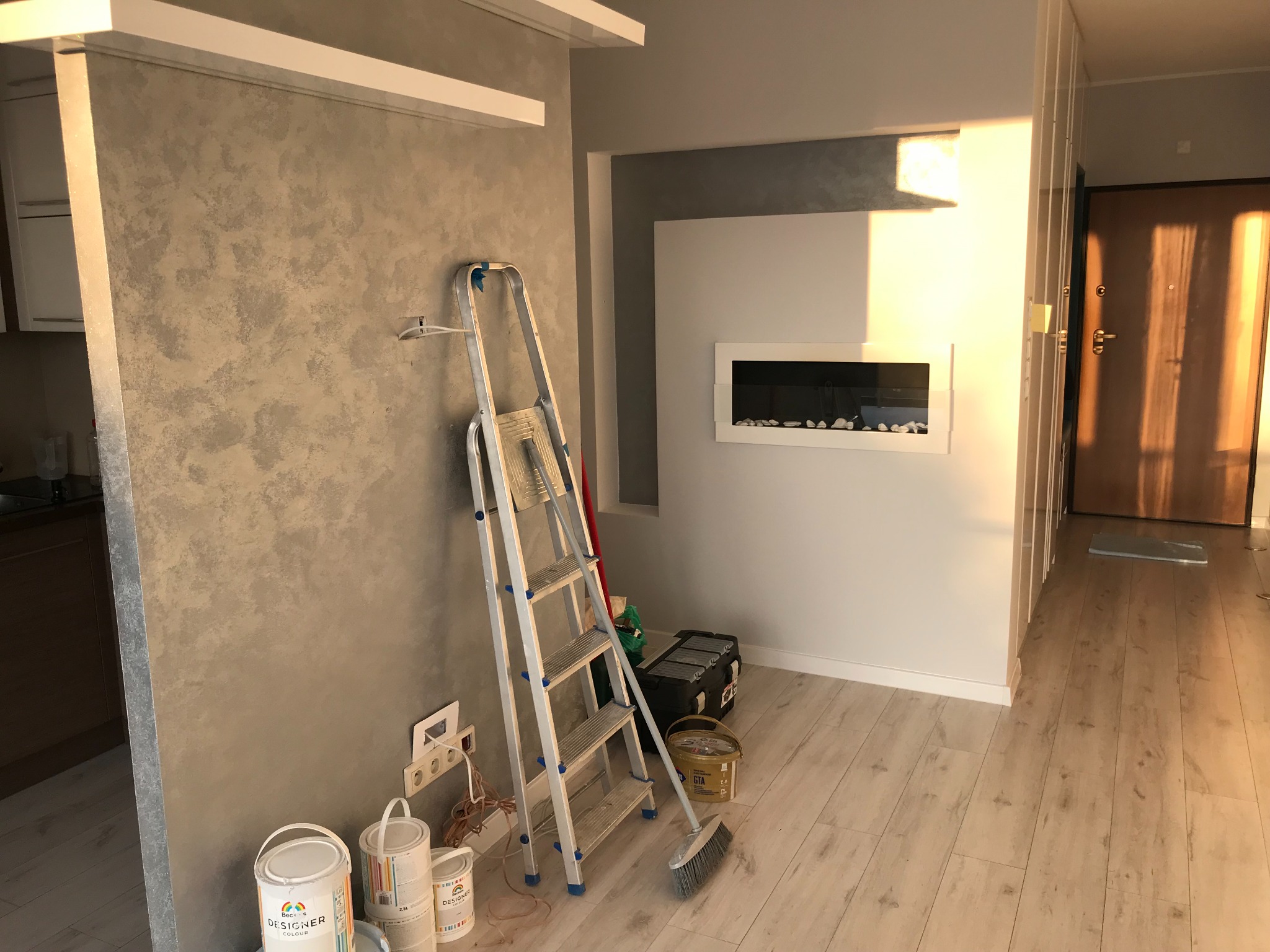 Ściana z efektem dekoracyjnym w odcieniach szarości, drabina malarska, puszki z farbą Designer Colour i narzędzia malarskie w jasnym wnętrzu z drewnianą podłogą i nowoczesnym kominkiem.