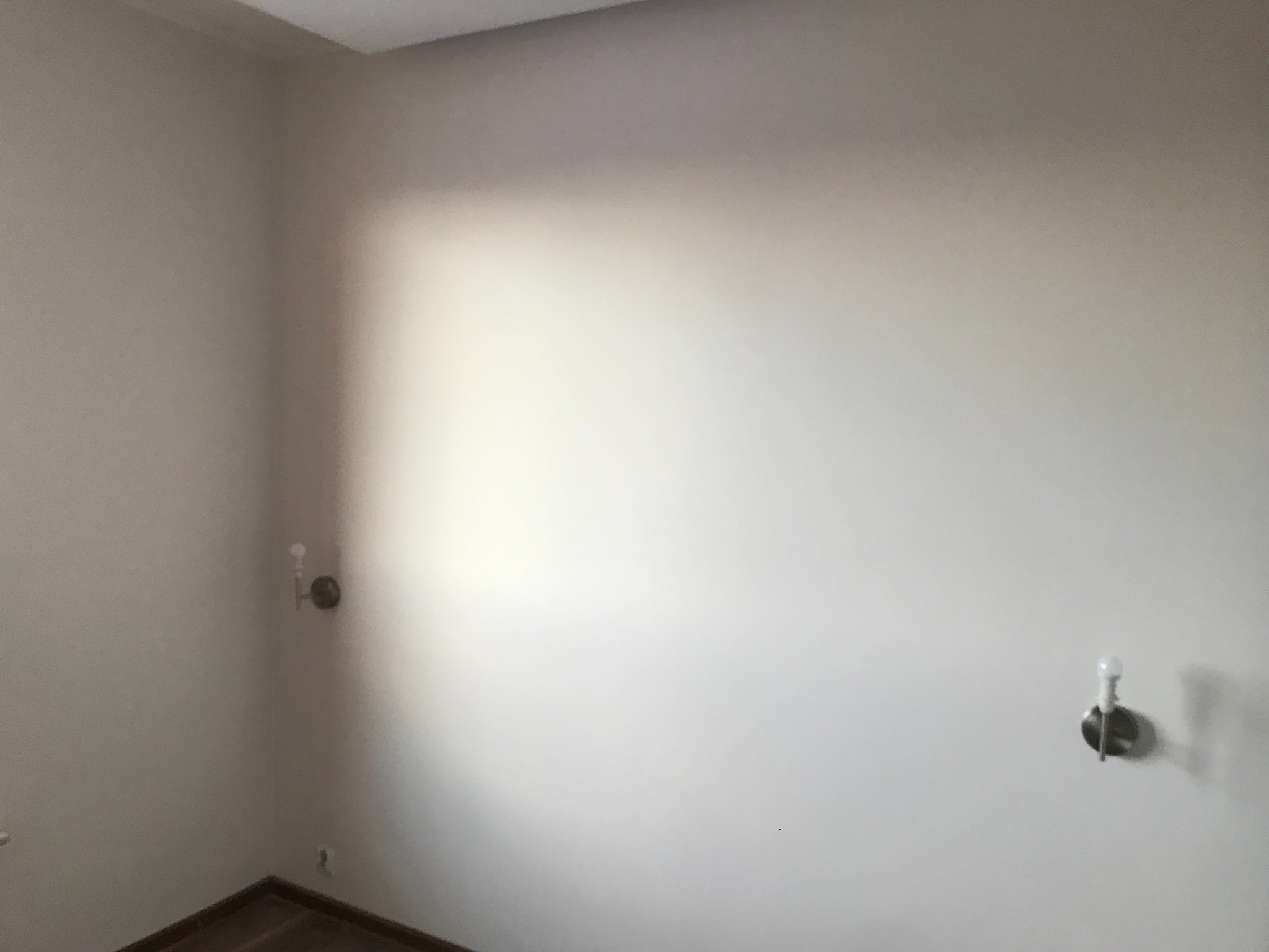 Pomalowane na jasny kolor ściany w rogu pokoju z dwoma kinkietami i fragmentem parkietu przy podłodze.