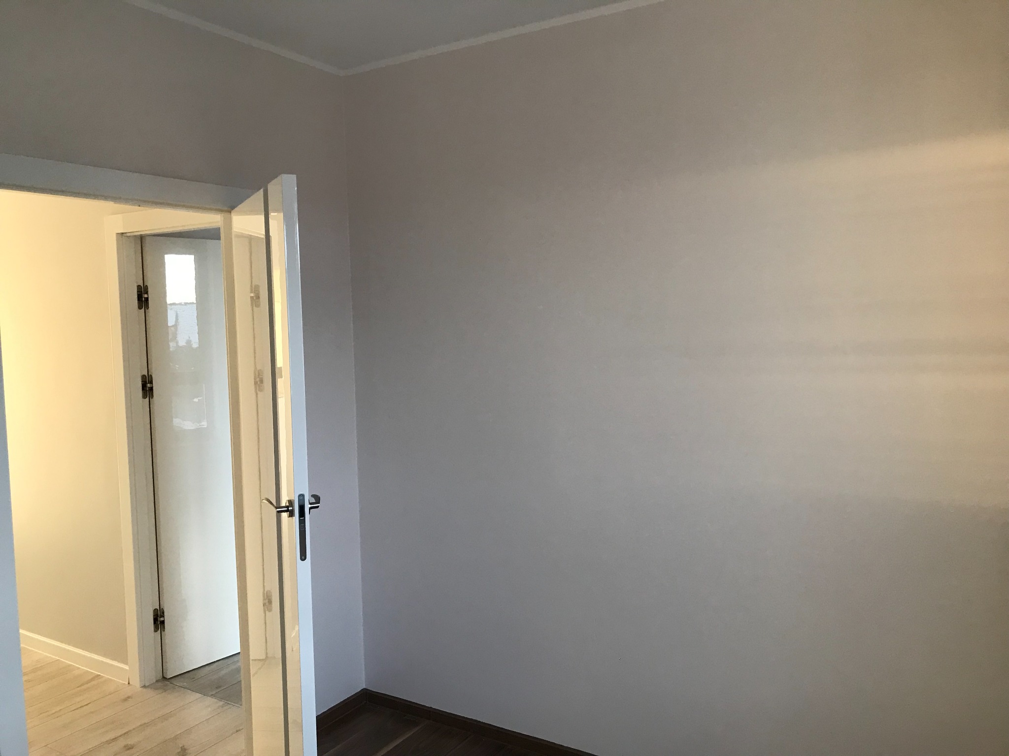 Pomalowane na jasny odcień szarości wnętrze pokoju z otwartymi białymi drzwiami, widoczne ciemne panele podłogowe.