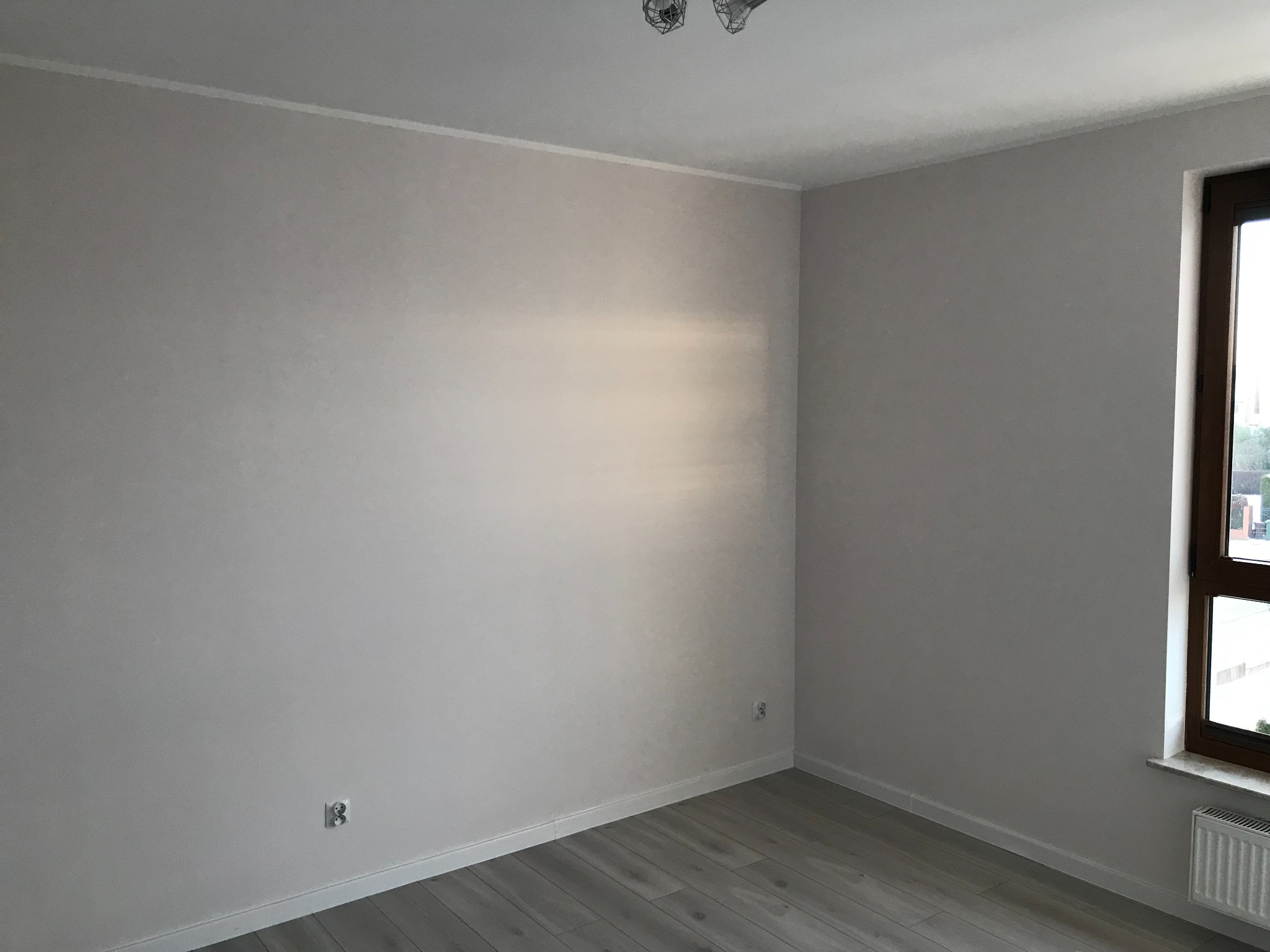 Pomalowane na jasny odcień szarości ściany w rogu pokoju z drewnopodobną podłogą, widoczne dwa gniazdka elektryczne i grzejnik pod oknem.