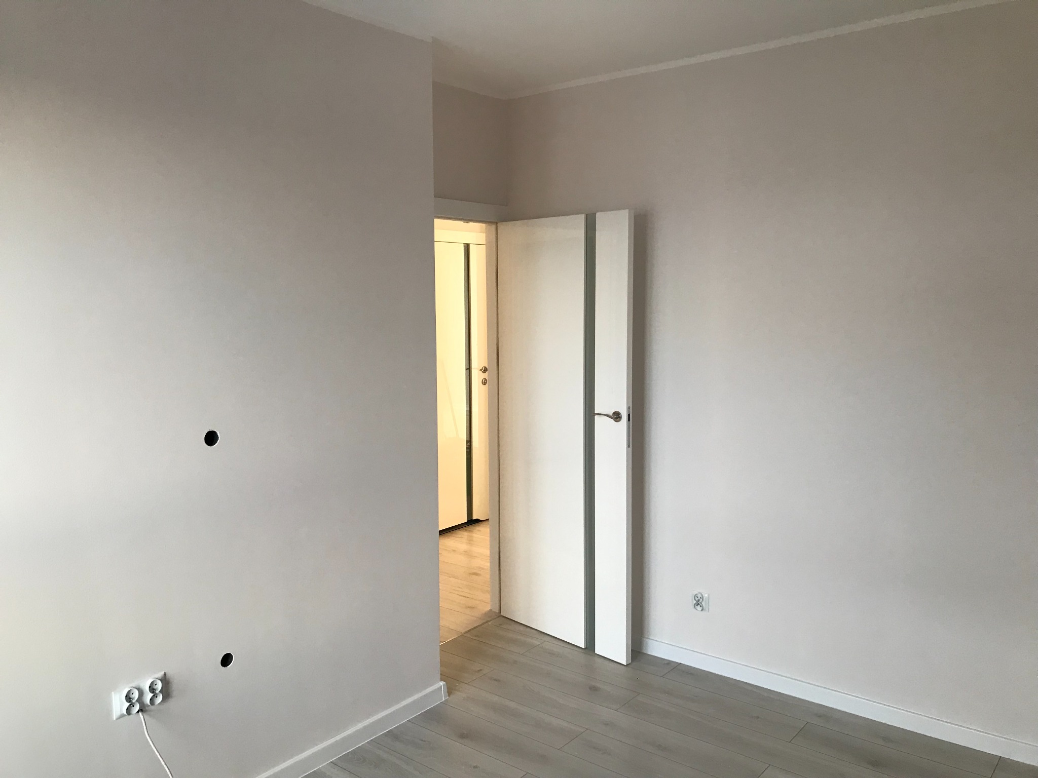 Świeżo pomalowane, jasnoszare ściany w pustym pokoju z otwartymi białymi drzwiami, widoczne gniazdka elektryczne i otwory w ścianie przygotowane do montażu osprzętu.