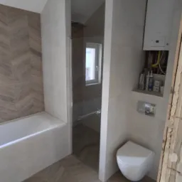 Jasna łazienka po remoncie z kabiną prysznicową, podwieszaną toaletą, grzejnikiem i płytkami ułożonymi w jodełkę na podłodze i ścianie przy wannie.