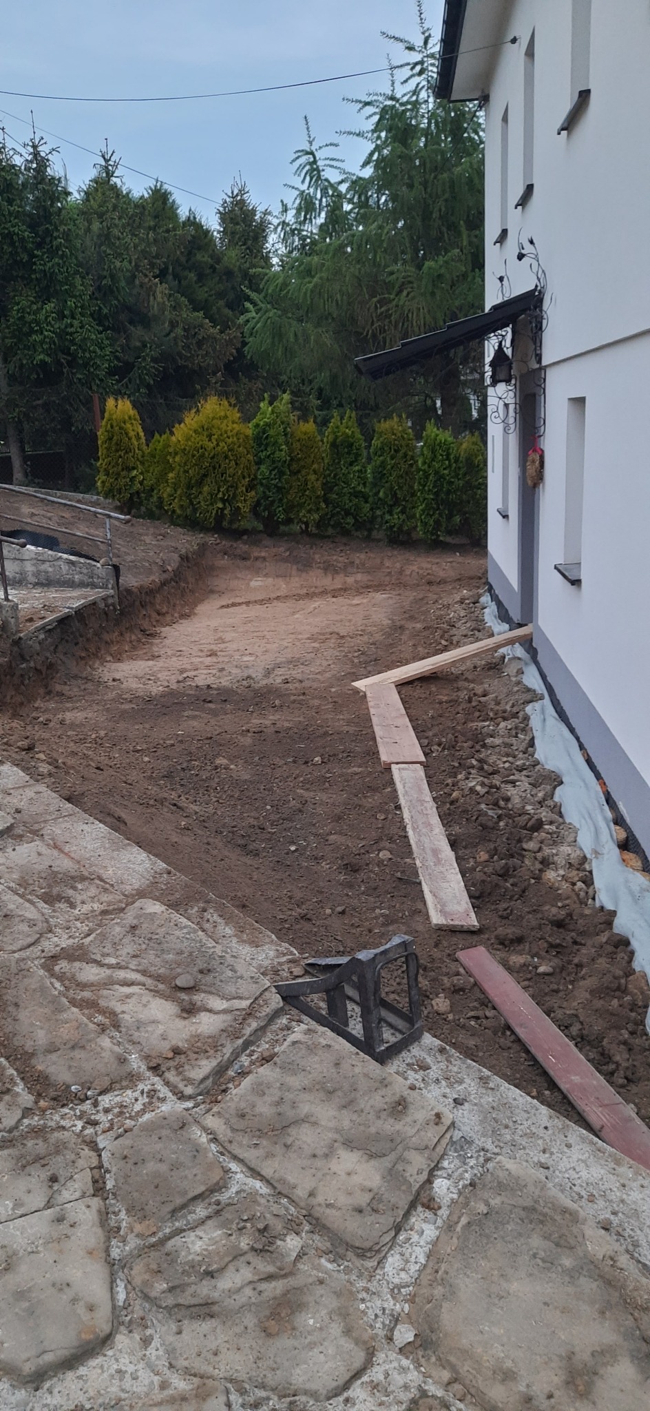 Przygotowany teren przy budynku z widocznym wykopem, zabezpieczeniem folią fundamentów, drewnianymi deskami i elementami kamiennej ścieżki.